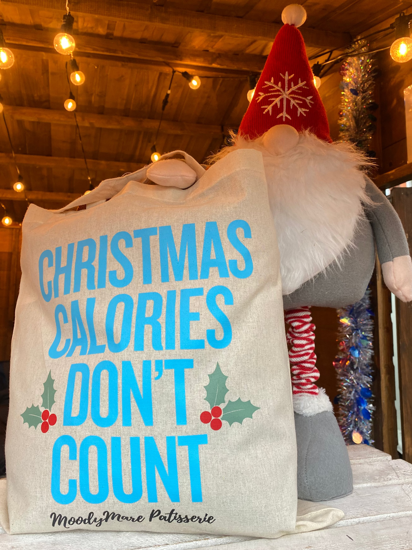 A large cream canvas tote bag with the text 'CHRISTMAS CALORIES DON'T COUNT' in bright blue, decorated with green holly and red berries, styled next to a Christmas gnome plush in a rustic indoor setting with warm string lights.